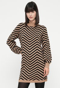 Kvinde med glat, mørkt hår iført en beige og sort chevron-mønstret langærmet sweaterkjole med puffede ærmer, stående mod en hvid baggrund.