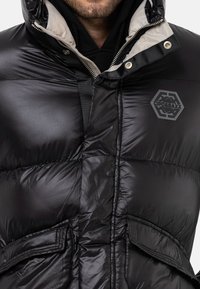 Schwarze Steppjacke mit glänzender Oberfläche, hohem Kragen, grauem Innenfutter, Reißverschluss vorne, Logo-Patch und zwei vorderen Taschen.
