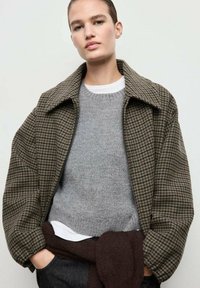 Chaqueta de cuadros marrón de gran tamaño con un gran collar, combinada con un suéter gris claro. Ajuste holgado y tejido texturizado, con acentos oscuros.