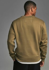Sudadera de color verde oliva con dobladillo y puños acanalados, textura suave y detalle de parche en la parte posterior. Manos posicionadas casualmente en las caderas.