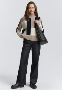 Pull à col roulé en beige, marine et blanc avec des motifs géométriques, associé à un pantalon en jean foncé évasé et des bottes noires à semelle épaisse.