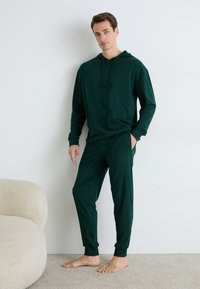 Grønn hettegenser og matchende joggebukser laget av mykt stoff, med en frontlomme og subtile logodetaljer på buksene. Teksturert overflate.