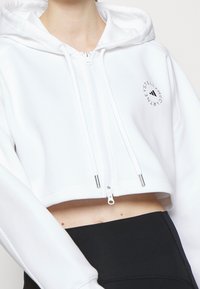 Sudadera corta blanca de tejido suave con cremallera frontal, cordones ajustables y un peque�ño detalle de logo negro en el pecho.