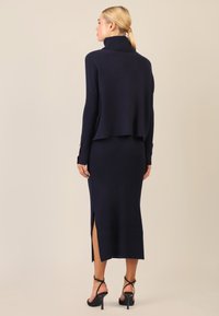 Navy-blaues geripptes Strickkleid mit Rollkragen, seitlichen Schlitzen und langen Ärmeln. Schwarze Sandalen mit Riemchen runden das Outfit ab.