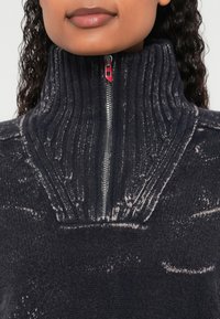 Tumši pelēks adīts džemperis ar ribotu augstu apkakli, sudraba rāvējslēdzēju un smalkām gaišākām rakstu detaļām audumā.