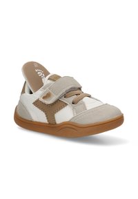 Zapato con tira de Velcro en material sintético blanco, con acentos en beige y suela de goma marrón. Presenta una puntera texturizada y detalles de costura.