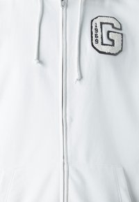 Bijela zip-up dukserica od glatkog materijala ima izvezeno crno-belo slovo "G" ispod kojeg se nalaze godine "1969". Uključuje i pertle.