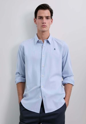 Camisa de botones color azul claro con mangas remangadas, sutiles rayas verticales y un pequeño logo bordado en el pecho. Textura de tela suave.