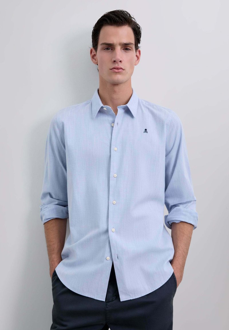 Camisa de botones color azul claro con mangas remangadas, sutiles rayas verticales y un pequeño logo bordado en el pecho. Textura de tela suave.