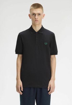 Joven con una camiseta polo negra de manga corta con un logo de laurel verde, de pie frente a un fondo liso gris claro.