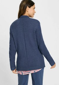 Navyblauwe cardigan met een textuur breiwerk, voorzien van een hoge kraag en lange mouwen. Aanzienlijk langere achterkant en ribgebreide boorden en zoom.