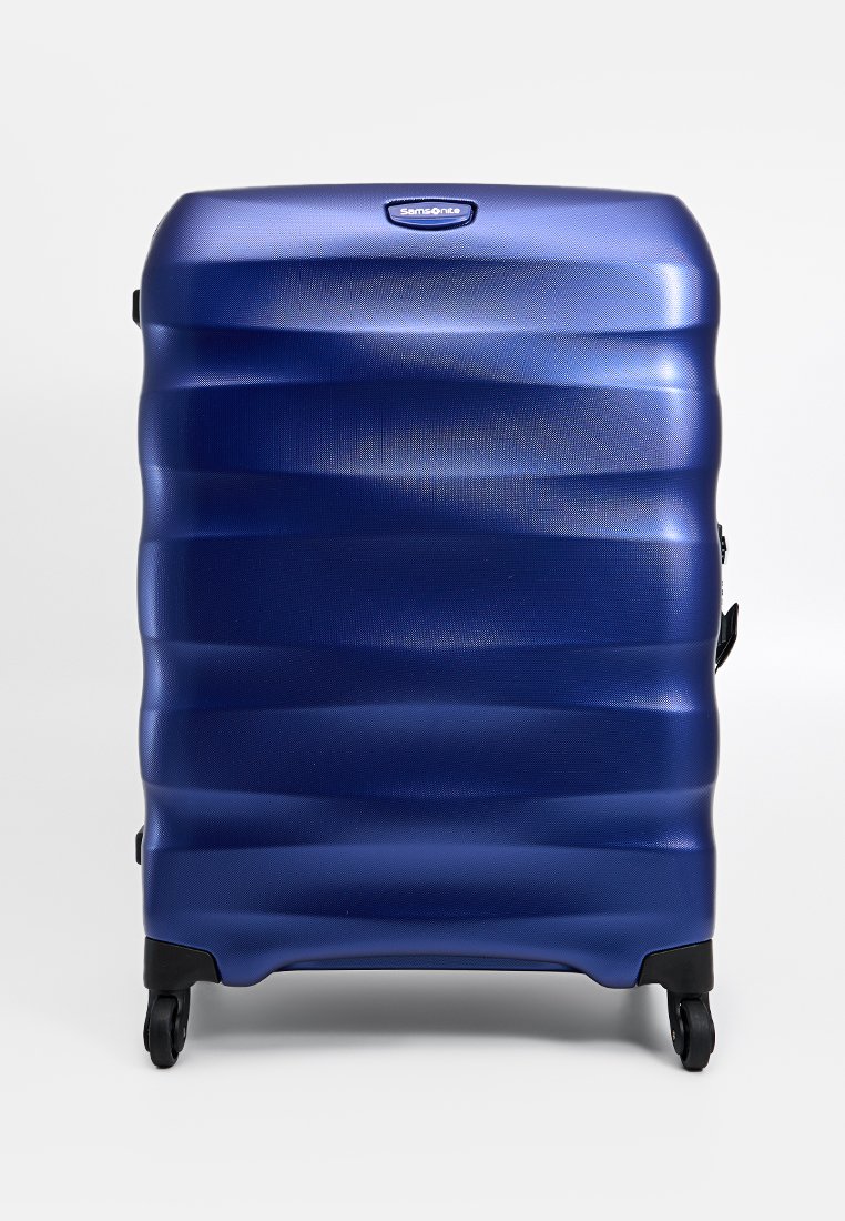 Valise rigide bleue avec un design à vagues texturé, équipée de quatre roues pour la mobilité et d'une poignée latérale pour un levage facile.