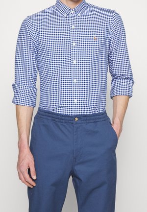 Mężczyzna ubrany w niebiesko-białą koszulę w kratę z podwiniętymi rękawami i granatowe spodnie z elastycznym pasem, jedna ręka w kieszeni.