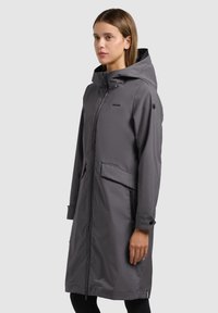 Mujer con un impermeable largo gris con capucha, cierre de cremallera y bolsillos con solapa, de pie frente a un fondo liso gris claro.