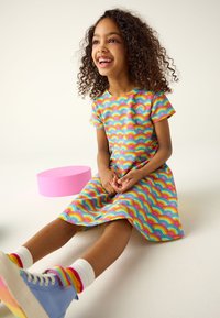 Obarvana obleka s pisanim vzorcem mavrice, s kratkimi rokavi, sproščenim krojem in okroglim izrezom. Kombinirana z modrimi supergami in pisano črtastimi nogavicami.