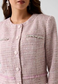 Veste en tweed rose pâle avec un tissage texturé, comprenant deux poches ornées d'accents métalliques en forme de feuille et une frange rose le long de l'ourlet.