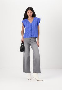 Blusa blu texturizzata con scollatura a V, maniche corte con rouches, bottoni frontali; abbinata a jeans grigi a gamba larga e stivali alla caviglia bianchi con tacco.