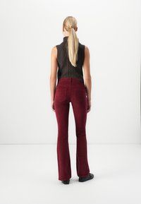 Femme avec une longue queue de cheval blonde portant un haut en tricot sans manches marron foncé et un pantalon en velours côtelé rouge taille haute, de dos sur fond blanc.