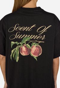 Zwarte t-shirt met een grafisch ontwerp van twee perziken met groene bladeren. Bovenaan staat de tekst "Scent Of Summer" in gouden script.