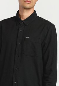 Chemise noire à boutons, en tissu texturé, avec une couleur unie, une poche poitrine et un petit logo sur la poche.