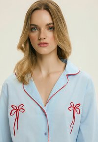 Młoda kobieta z blond włosami do ramion nosi jasnoniebieską koszulę z czerwonymi haftowanymi kokardkami oraz czerwonym wykończeniem na kołnierzyku i guzikach.