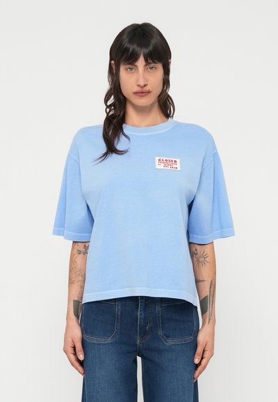 T-shirt à manches courtes bleu clair avec une coupe carrée, arborant une étiquette de logo rouge et blanche sur le côté gauche de la poitrine. Matière en coton avec une texture lisse.