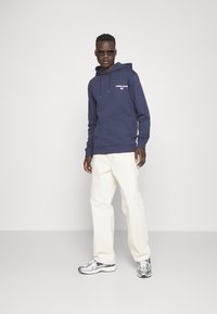 Felpa blu navy con logo bianco "TOMMY JEANS", abbinata a pantaloni bianchi e sneaker argento. Il modello indossa anche occhiali da sole scuri.