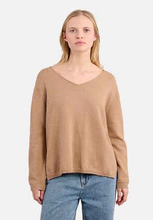 Donna con capelli biondi lisci che indossa un maglione a V di colore marrone chiaro e pantaloni jeans blu, su uno sfondo uniforme.