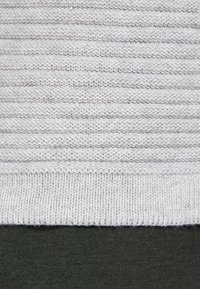 Ljusgrå stickad tröja med ett strukturerat randigt mönster. Ribbad nederdel och ärmslut, tillverkad av mjukt material. Bärs över ett mörkgrönt plagg.