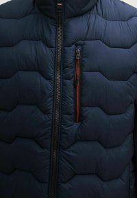 Veste en duvet marine avec un design matelassé, fermeture éclair noire et une poche poitrine avec un détail de fermeture éclair rouge. Tissu texturé.