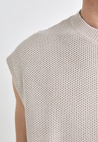 Lys beige, ermeløs strikket topp med en strukturert mønster og ribbestrikket halslinning, som viser en tettsittende design.