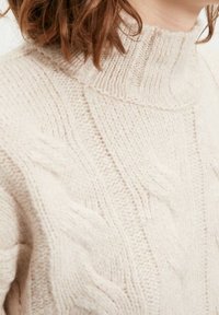 Pull beige en maille avec un col montant, des torsades texturées et des accents côtelés. Le fil doux et épais offre un aspect chaleureux.