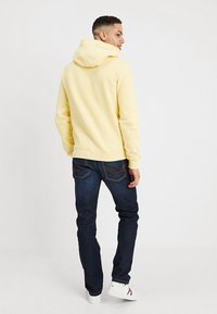 Geel hoodie met een trekkoordcapuchon, gecombineerd met donkerblauwe jeans. De outfit heeft een ontspannen pasvorm en textuur, afgebeeld vanaf de achterkant.