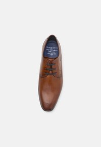 Chaussure en cuir marron avec un design perforé, bout arrondi et lacets noirs. L'intérieur présente une étiquette de marque et une semelle intérieure rembourrée.