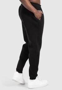 Černé bavlněné tepláky s tapered nohavicemi a elastickým pasem. Obsahují boční kapsy a žebrované manžety. Vybavené bílými teniskami.