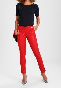 Kobieta w czarnej bluzce z krótkim rękawem, czerwonych slim-fit spodniach, srebrnych szpilkach i nosząca czarną i czerwoną torebkę na ramię.