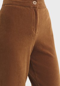 Brown corduroy pants with vertical rib texture, featuring a front button closure and side pocket. The fabric appears soft and durable.