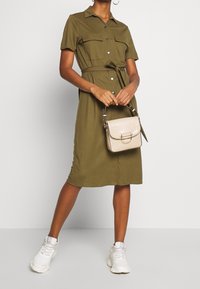 Abito verde oliva con cintura, maniche corte, apertura con bottoni, due tasche sul petto, abbinato a una borsa beige e scarpe da ginnastica bianche.