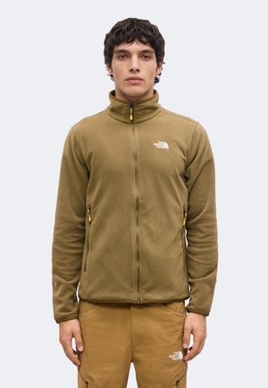 Junger Mann trägt eine braune Fleecejacke mit Reißverschluss und passende Hose mit The North Face Logos, stehend vor einfachem weißem Hintergrund.