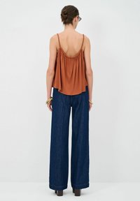 Top senza maniche color ruggine con spalline intrecciate e design arricciato, abbinato a pantaloni in denim scuro a gamba larga, visto da dietro.