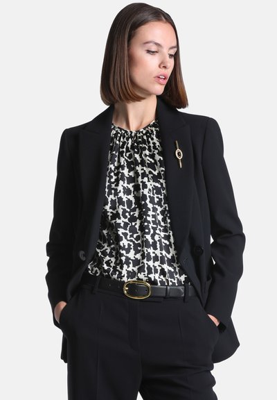 Femme portant un blazer noir avec une broche, un chemisier à motif noir et blanc, une ceinture noire et un pantalon noir, les mains dans les poches.