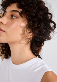 Orecchino a cerchio in oro con un design intrecciato. Il modello ha i capelli ricci e indossa una canotta bianca. La luce soffusa evidenzia la texture.