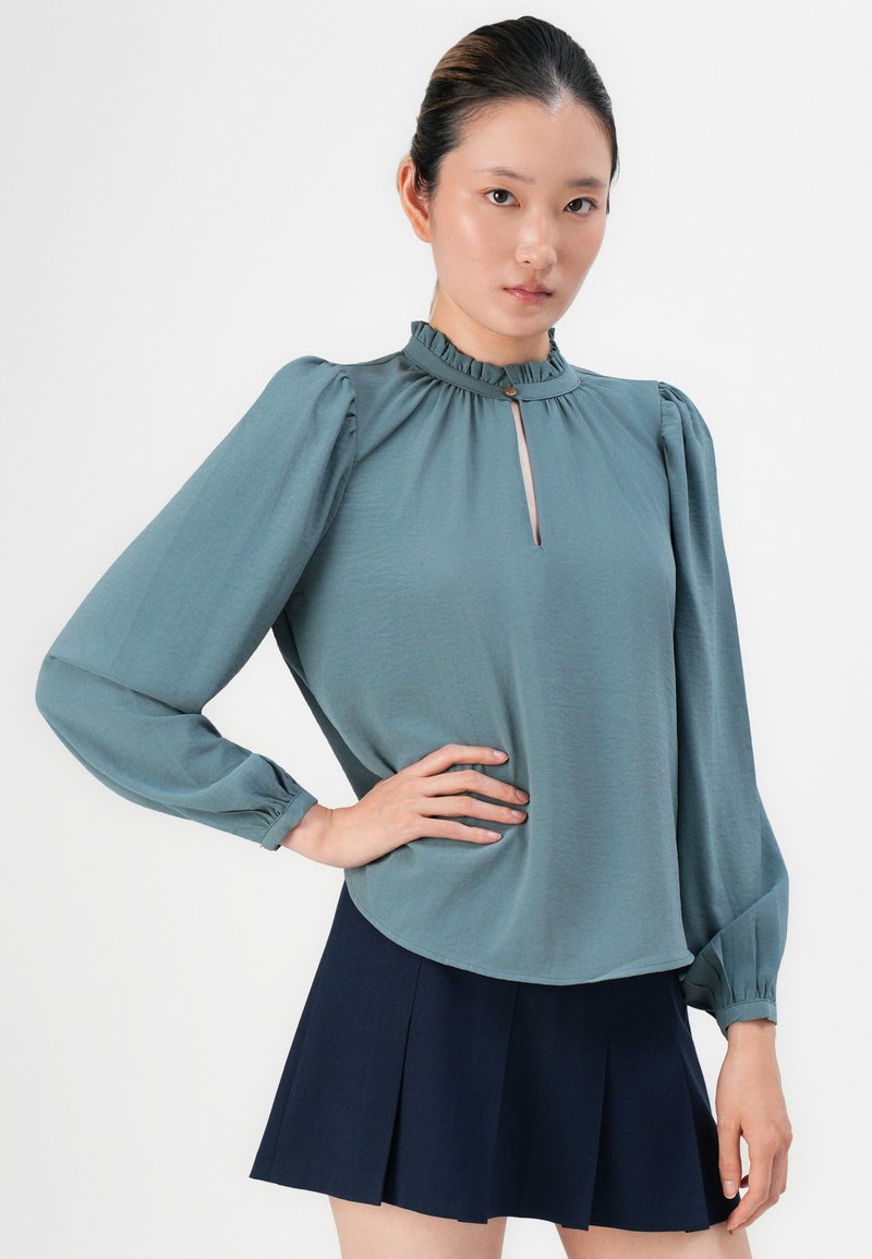 Teal langemouwtop met een ruches kraag en keyhole detail bij de halslijn, gecombineerd met een marineblauwe geplooide rok. Zachte stoftextuur.