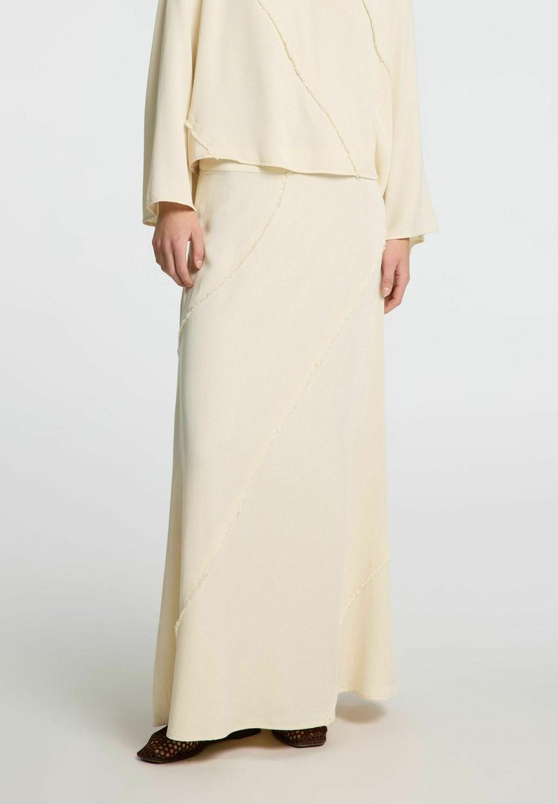 Conjunto de dos piezas beige que incluye una falda larga y fluida y una blusa holgada. Tejido suave con detalles de costura diagonal y bordes deshilachados.