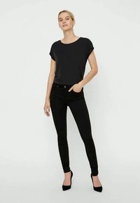 Črna majica s kratkimi rokavi v kombinaciji s črnimi skinny jeans. Model nosi črne visoke pete. Enostaven dizajn, gladka tekstura in oprijet kroj.