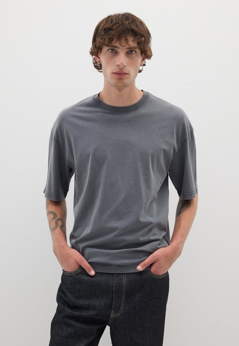 T-shirt grigio a maniche corte realizzato in tessuto morbido, con colletto a girocollo e vestibilità rilassata. Abbinato a jeans in denim scuri con dettagli di cucitura.