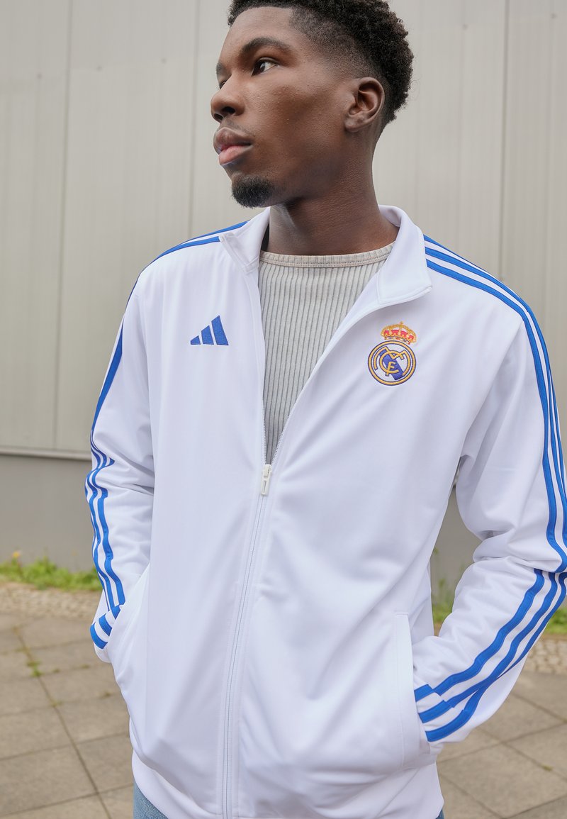 Hvit jakke med glidelås og blå Adidas-striper samt Real Madrid-logo. Har en ribbet grå krage og frontlommer. Glatt stoffstruktur.