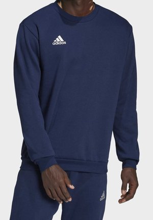 Mężczyzna ubrany w granatową bluzę Adidas z okrągłym dekoltem i pasujące spodnie dresowe, z widocznym logo na piersi i udzie.