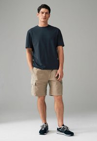 Donkergrijze katoenen t-shirt voor mannen, beige cargo shorts met zijzakken, en zwarte sportschoenen met grijze accenten. Eenvoudige, losse pasvorm.
