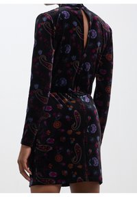 Femme portant une robe en velours noire à manches longues avec des motifs floraux et cachemire colorés, vue de dos avec un détail d’encolure en forme de trou de serrure.